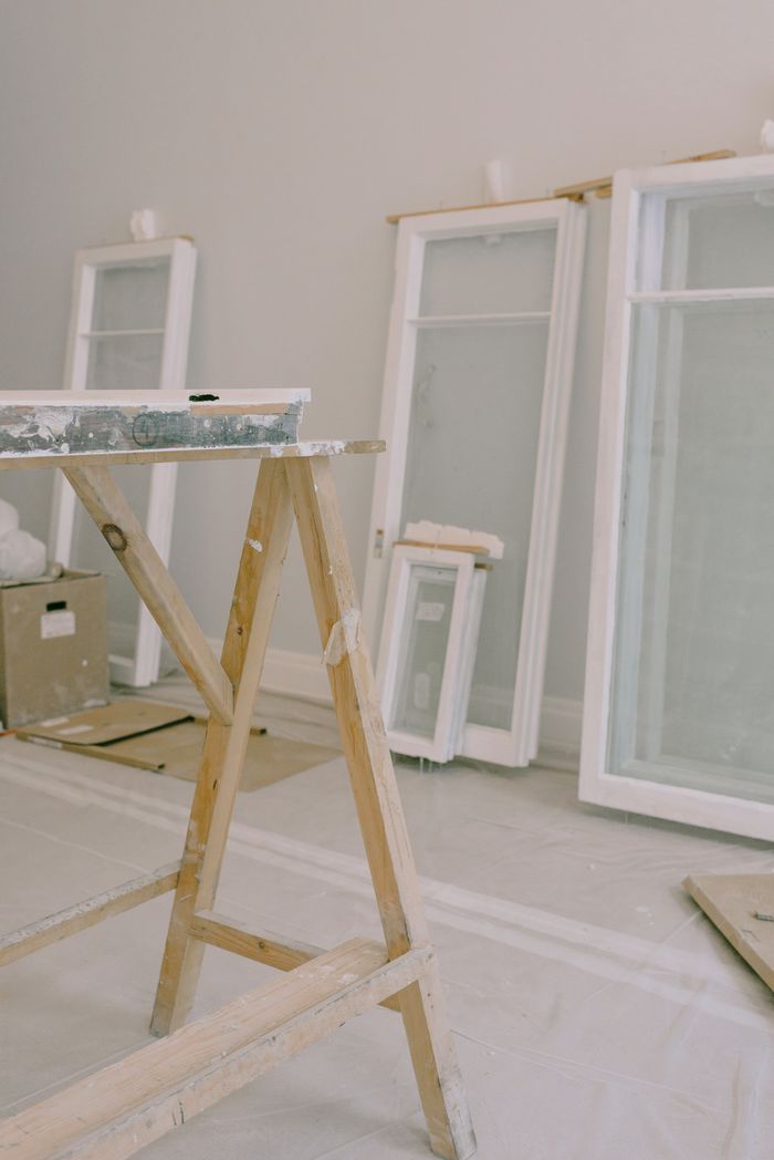 Interior of a room featuring a wooden workbench and windows taken off their hinges, with a white frame set beside the wall in a bright room during repair work